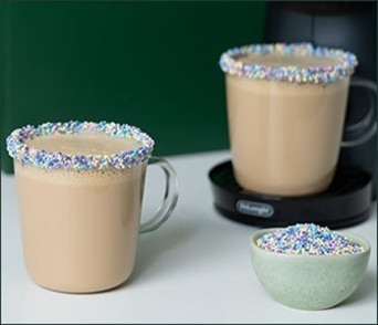 Twee mokken romige koffie met randen versierd met kleurrijke sprinkles en een klein bakje sprinkles